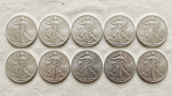 Ten silver-colored coins arranged in two rows of five on a light beige textured fabric background.