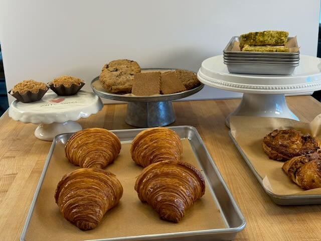 Artisan bakery spread with golden butter croissants on sheet pan muffins cookies and layered pastry bars on white cake stands and baking trays