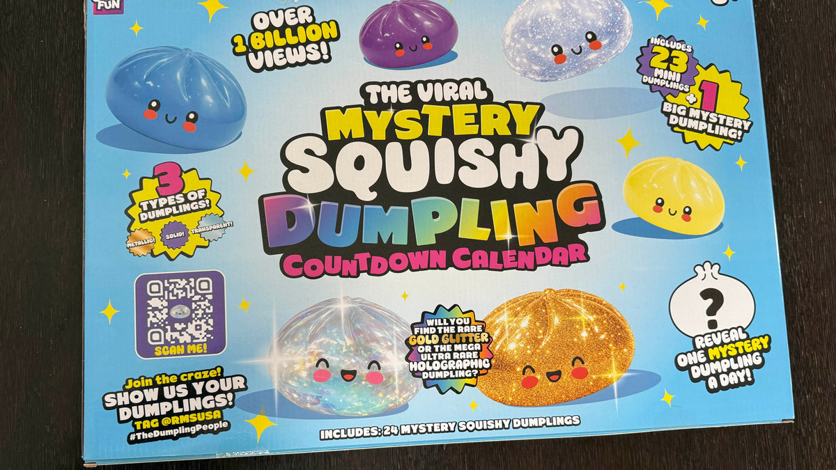 The Viral Mystery Squishy Dumpling Countdown Calendar toy box on a dark wood background. The box features a bright blue background with cartoonish squishy dumplings in various colors (blue, purple, clear, yellow, orange) and colorful text advertising the product.