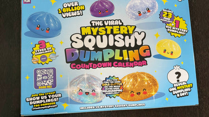 The Viral Mystery Squishy Dumpling Countdown Calendar toy box on a dark wood background. The box features a bright blue background with cartoonish squishy dumplings in various colors (blue, purple, clear, yellow, orange) and colorful text advertising the product.