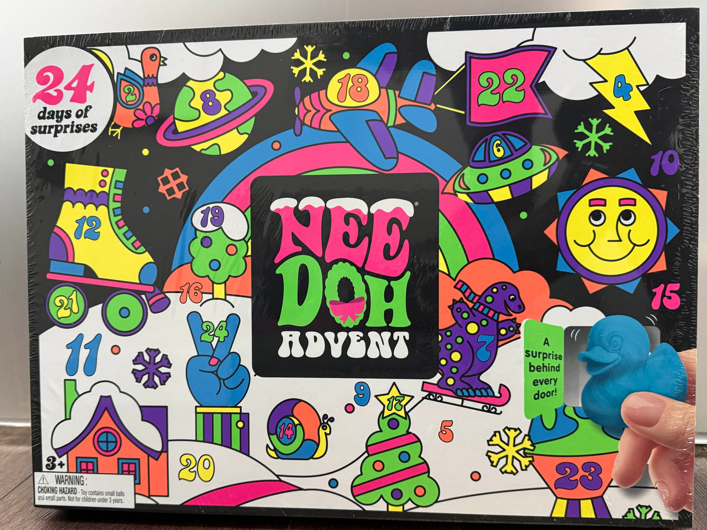 A colorful NEE DOH Advent box with a hand opening one of the numbered surprise doors. The box features a vibrant, cartoon-style illustration with 24 days of surprises text and a blue dinosaur surprise peeking out.