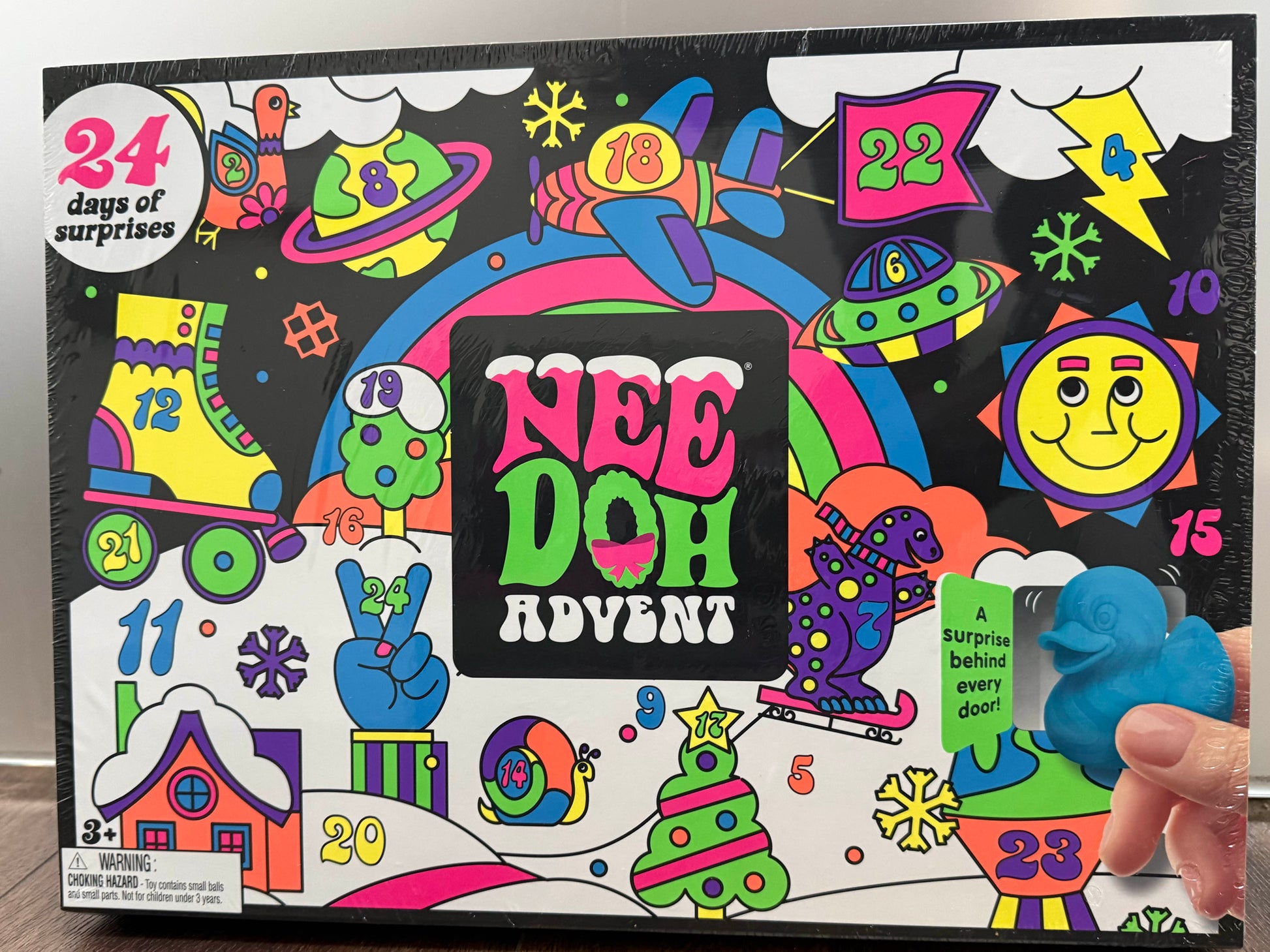 A colorful NEE DOH Advent box with a hand opening one of the numbered surprise doors. The box features a vibrant, cartoon-style illustration with 24 days of surprises text and a blue dinosaur surprise peeking out.